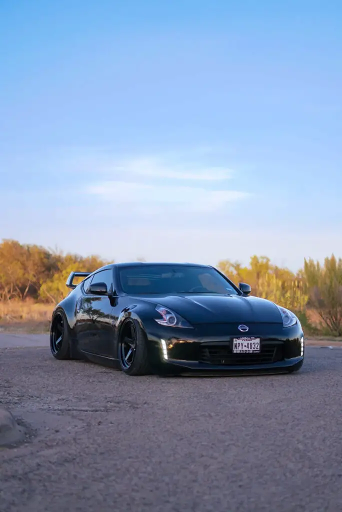 A black Nissan 370Z with aftermarket wheels and tires, parked on a street with a clear blue sky in the background.